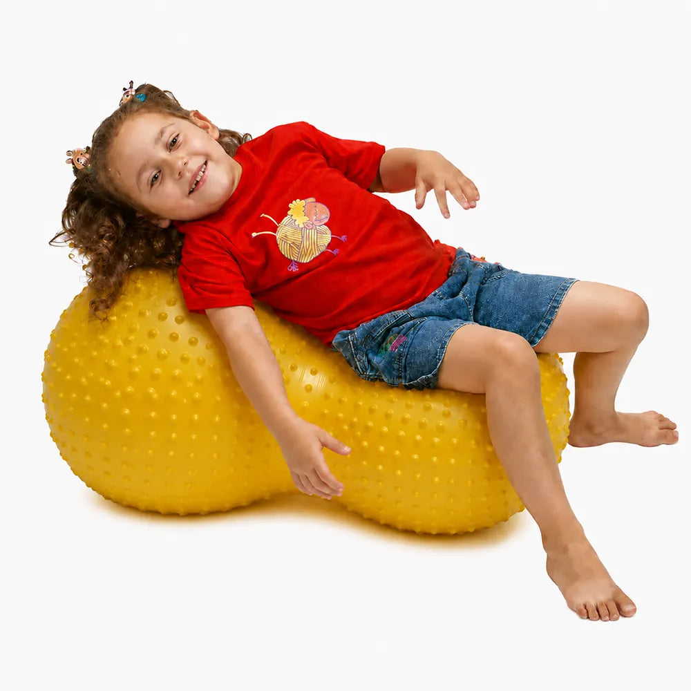 Enfant assise sur un ballon sensoriel jaune à picots en forme de cacahuète.